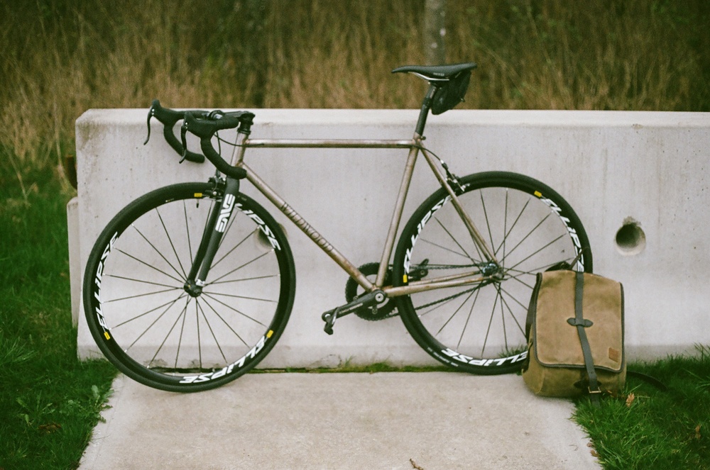 Wildwood Bicyles - a custom bicycle leans against a concrete block with tall grass behind it, a saddle bag on the ground by its back wheel.